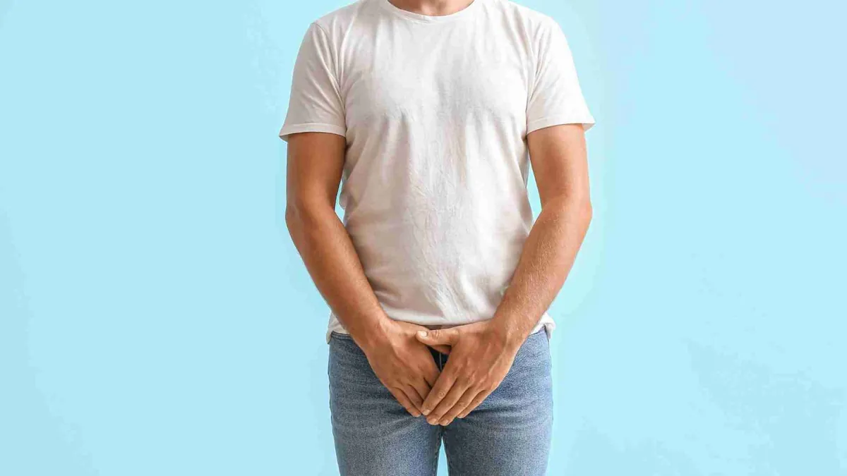 Hombre con camiseta blanca y las manos sobre la zona pélvica frente a un fondo azul